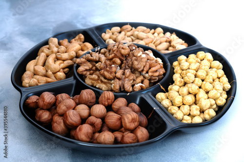 A round black serving platter with five compartments full of assorted shelled nuts (hazelnuts, cashews, peanuts, walnuts, and chickpeas) sits on a light gray table. Close-up