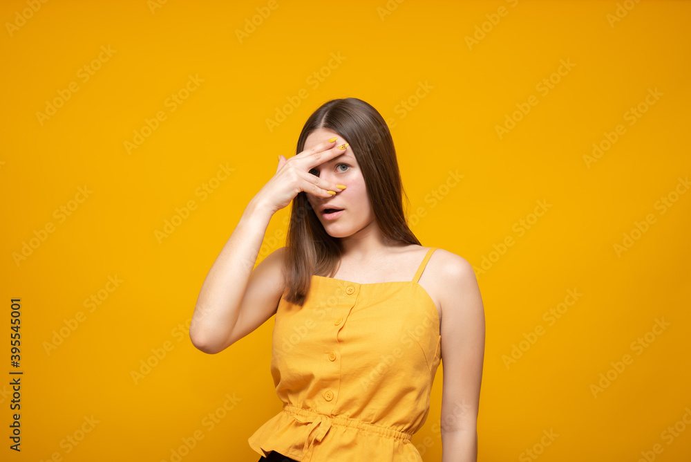 Young beautiful caucasian woman in a yellow sweater on a yellow background in shock, covering her face and eyes with her hand, looking through her fingers with an embarrassed expression. Young beautiful caucasian woman in a yellow sweater on a yellow background in shock, covering her face and eyes with her hand, looking through her fingers with an embarrassed expression.