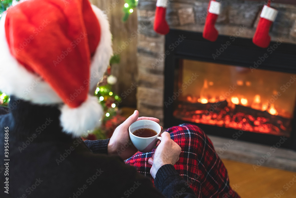 Obraz premium One divorce man with cup coffee, tea in santa claus hat sitting and warming at winter evening near fireplace flame, covered christmas plaid