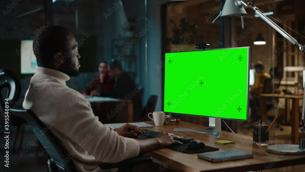 Handsome Black African American Project Manager is Making a Video Call on Desktop Computer with Green Screen Mock Up Display in a Busy Creative Office. Male Specialist is Wearing Turtle Neck Sweater. Handsome Black African American Project Manager is Making a Video Call on Desktop Computer with Green Screen Mock Up Display in a Busy Creative Office. Male Specialist is Wearing Turtle Neck Sweater.