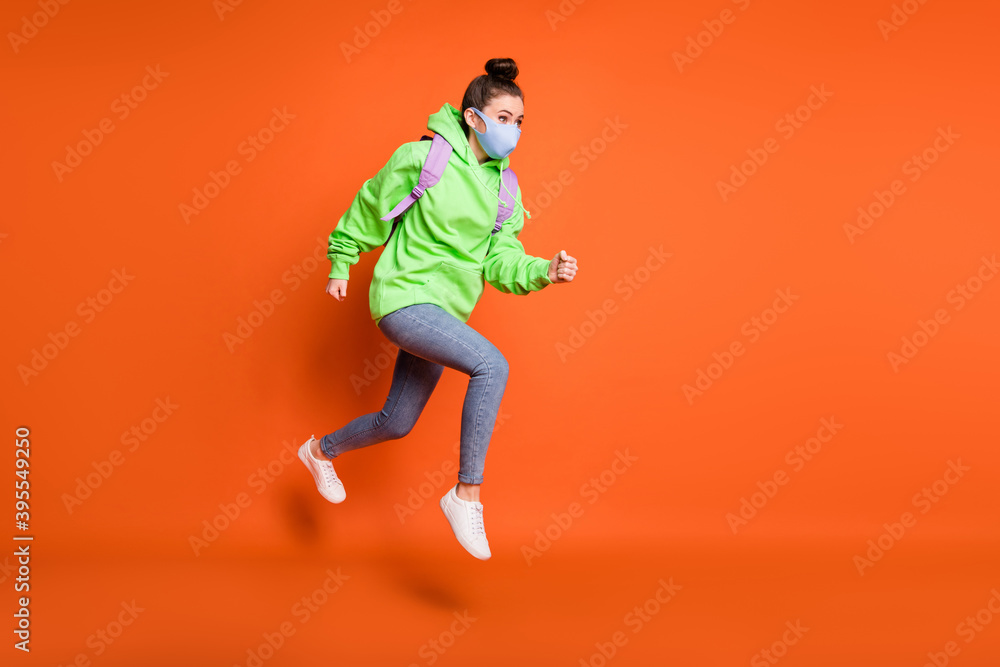 Photo portrait full body view of young girl jumping up running wearing blue fabric face mask isolated on vivid orange colored background Photo portrait full body view of young girl jumping up running wearing blue fabric face mask isolated on vivid orange colored background
