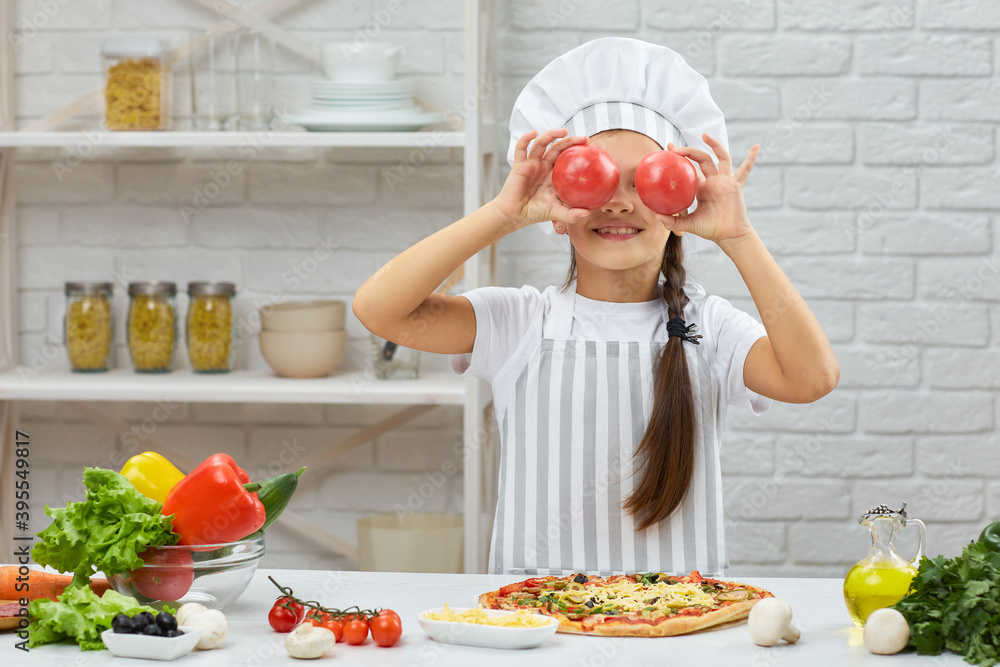 the child holding red tomato. little girl in chef hat and an apron cooking pizza in the kitchen. the child holding red tomato. little girl in chef hat and an apron cooking pizza in the kitchen.