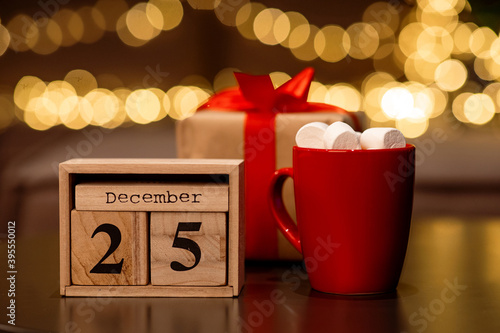 Cocoa with marshmallows, wooden December 25 calendar and a gift with a red ribbon on a table against a background with lights in bokeh.