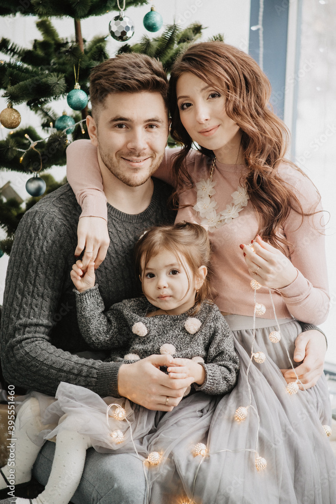 Young family with a daughter in festive outfits with a garland near the Christmas tree on New Years Eve Young family with a daughter in festive outfits with a garland near the Christmas tree on New Years Eve