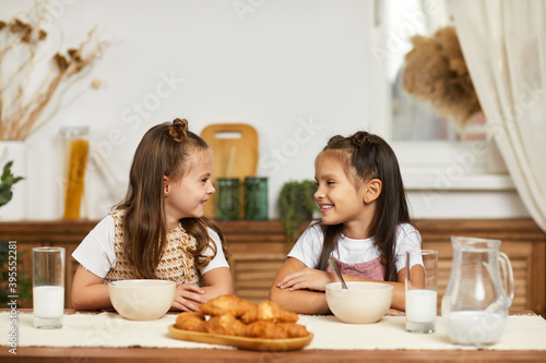 Cute healthy little children girls having breakfast - fresh delicious croissants and milk in the kitchen.