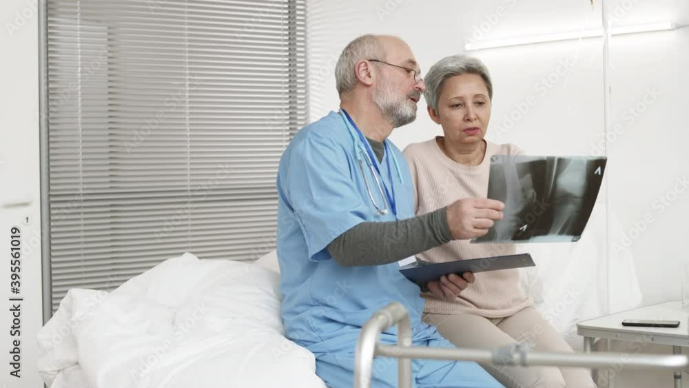 Medium shot of senior Asian woman and aged Caucasian doctor wearing blue uniform sitting on bed in hospital ward and analyzing x-ray picture Medium shot of senior Asian woman and aged Caucasian doctor wearing blue uniform sitting on bed in hospital ward and analyzing x-ray picture