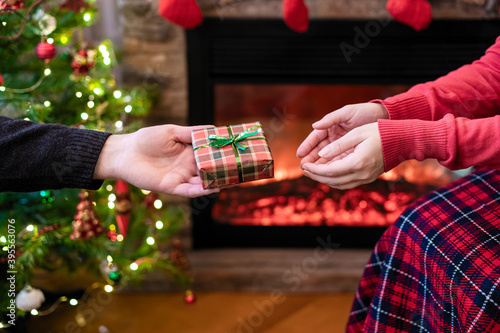 Man giving Christmas and New Year Gift box to woman in front of christmas tree, fireplace with flame. Xmas concept.