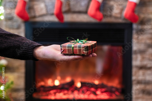 Fototapeta Naklejka Na Ścianę i Meble -  Man giving Christmas and New Year Gift box to woman in front of christmas tree, fireplace with flame. Xmas concept.
