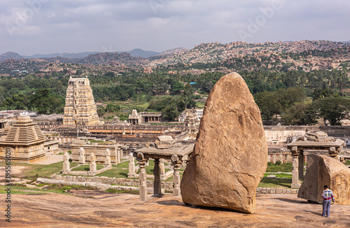 Hampi, Karnataka, India - November 4, 2013: Virupaksha Temple complex. Wide view over temple grounds with up front giant brown boulder on plateau above. Green forested area, brown hills and light blue photo