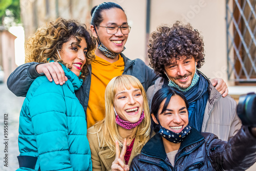 smiling happy group of friends taking a selfie portrait with a reflex camera wearing warm winter clothes and protective face mask to celebrate coronavirus lockdown reopening. millennial group people