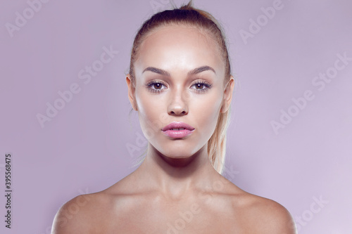 Front closeup portrait of a blonde beautiful girl with ponytail hairstyle and bare shoulders, on purple background.
