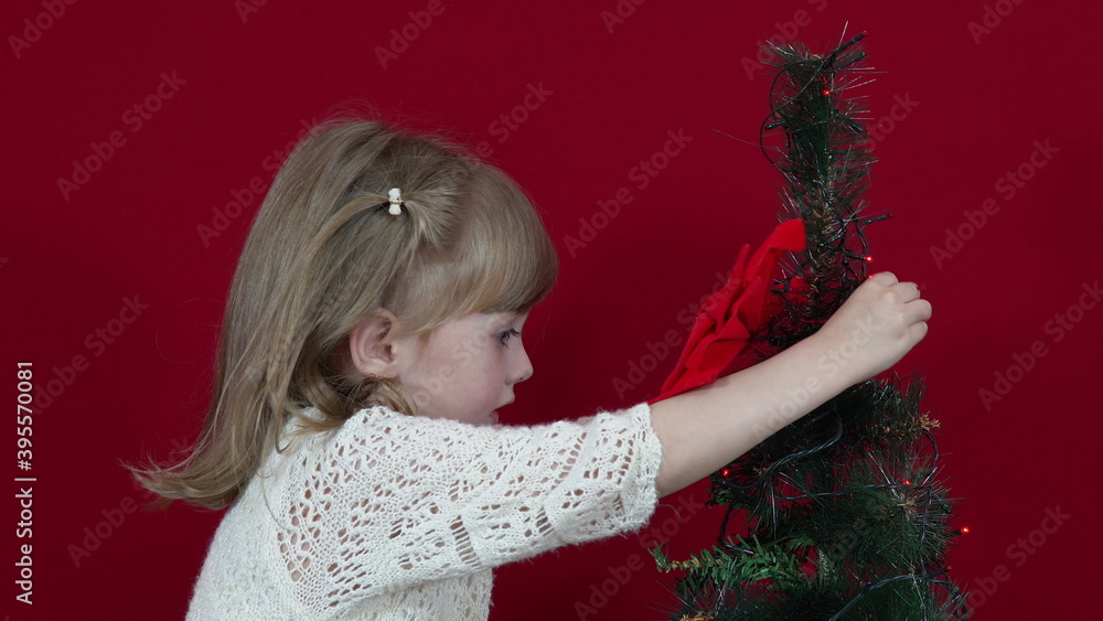 A girl in an elegant dress on a red background decorates a Christmas tree