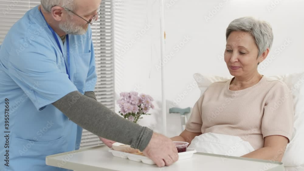 Medium shot of Caucasian male doctor bringing food tray to aged female patient lying in bed in hospital. Woman having lunch in hospital Medium shot of Caucasian male doctor bringing food tray to aged female patient lying in bed in hospital. Woman having lunch in hospital
