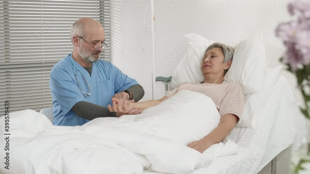 Medium shot of Caucasian physician wearing blue overall sitting on chair near bed and measuring pulse of ill old Asian woman lying on it Medium shot of Caucasian physician wearing blue overall sitting on chair near bed and measuring pulse of ill old Asian woman lying on it