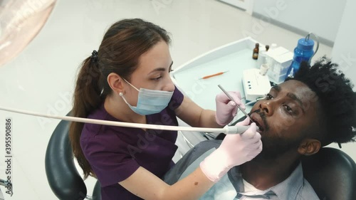 Professional female dentist in bluewhite protective gloves and violet medical suit, making examination and treatment of teeth of young handsome african male patient, in modern dental clinic. photo