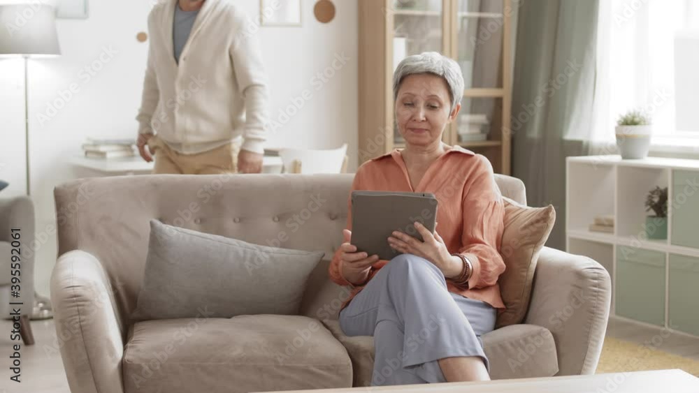 Medium shot of aged Caucasian man sitting at desk in living room, using laptop then coming to his wife sitting on sofa with tablet in her hands. They choosing new furniture online Medium shot of aged Caucasian man sitting at desk in living room, using laptop then coming to his wife sitting on sofa with tablet in her hands. They choosing new furniture online