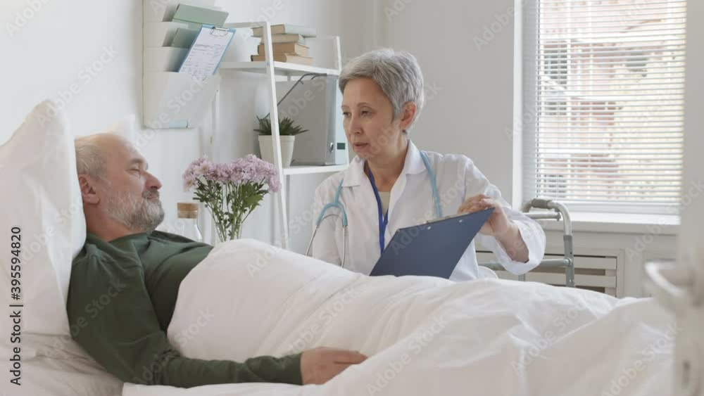 Medium shot of sick old Caucasian man lying in bed in hospital room and listening to female aged doctor with patient card in hands sitting nearby and speaking Medium shot of sick old Caucasian man lying in bed in hospital room and listening to female aged doctor with patient card in hands sitting nearby and speaking