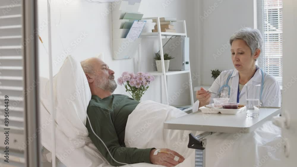 Medium shot of senior Asian female therapist sitting at bed in hospital ward and feeding Caucasian man lying in it Medium shot of senior Asian female therapist sitting at bed in hospital ward and feeding Caucasian man lying in it