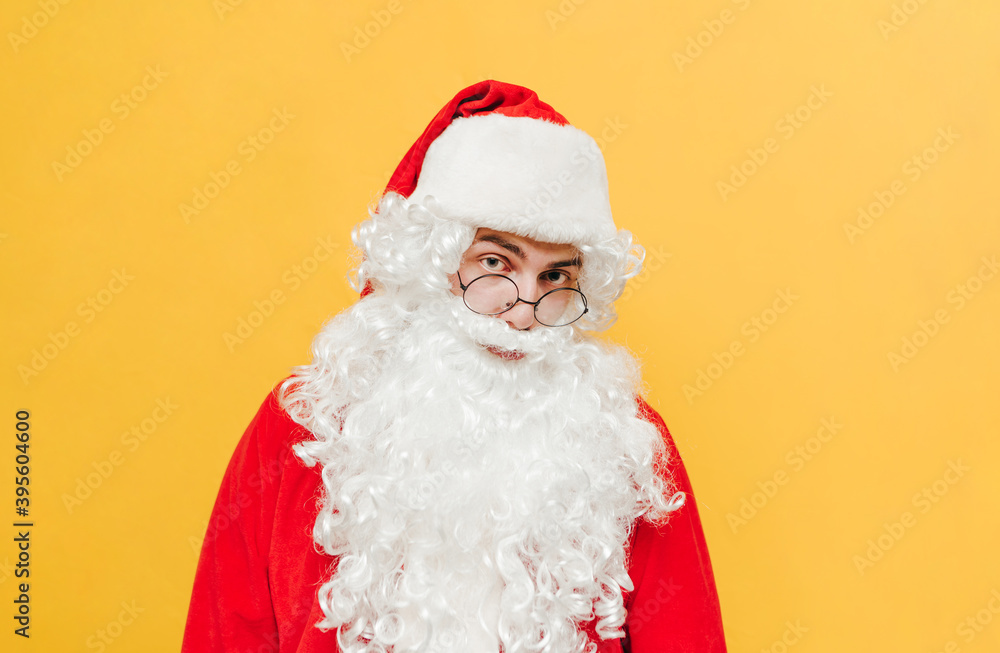 Closeup portrait of a Santa Claus standing at the studio on a yellow background, looking at the camera with an interested expression on his face with eyeglasses on his nose.