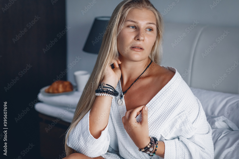 Beautiful blonde girl sitting on a bed with white linens in a hotel room in a white bathrobe after spa procedures Beautiful blonde girl sitting on a bed with white linens in a hotel room in a white bathrobe after spa procedures