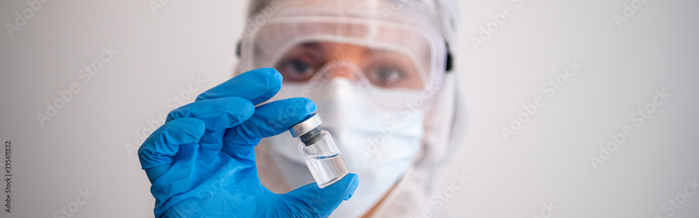 Panoramic - Female doctor wearing infection protective gear against coronavirus covid-19 virus, holds a medical vial full of liquid. Panoramic - Female doctor wearing infection protective gear against coronavirus covid-19 virus, holds a medical vial full of liquid.