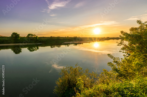 Fototapeta Naklejka Na Ścianę i Meble -  Scenic view at beautiful summer river sunset with reflection on water with green bushes, grass, golden sun rays, calm water ,deep blue cloudy sky and glow on a background, spring evening landscape