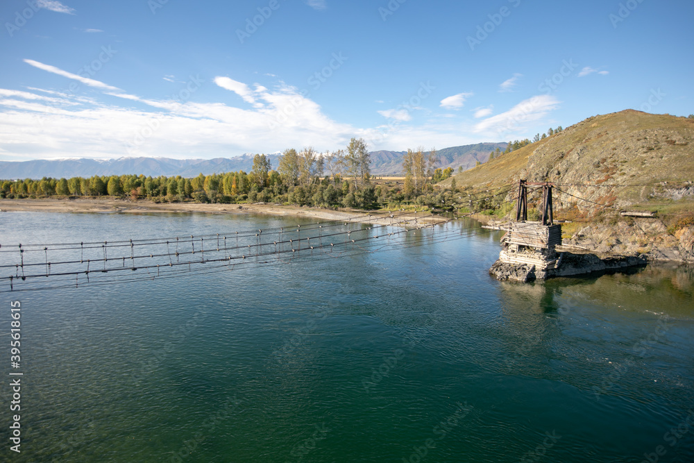 Fototapeta premium The very old hanging footbridge across river.Beautiful mountain landscape, blue river and coniferous forest