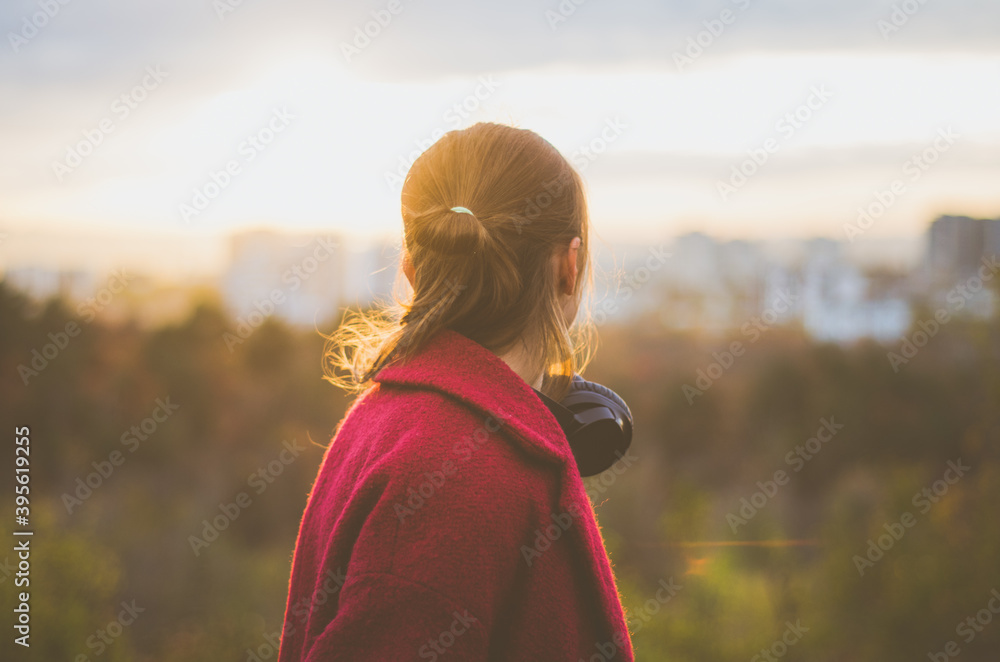 Back view portrait of a young caucasian woman with headphones around neck, looking serious to the sky, with blurred city on the background. Concept: leisure activity, mental health and well being