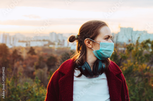 Side view of a woman wearing a face mask and headphones around neck walking the park at sunset. Concept: safety during the covid 19 pandemic, mental health and well being