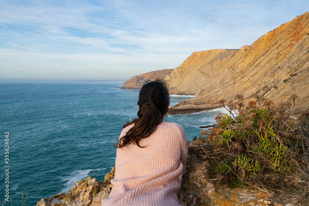 Woman on a pink shirt looking at the landscape of Cabo Espichel Cape sea cliffs and atlantic ocean, in Portugal