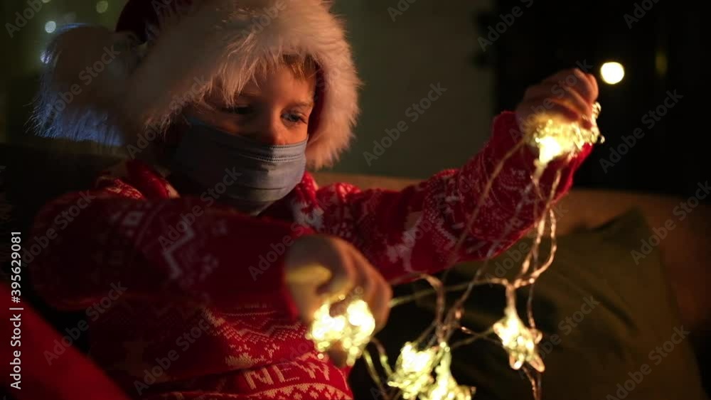 Christmas kids mask. Boy in Santa Claus hat decorates room with christmas lights, preparing for the holiday at home Christmas kids mask. Boy in Santa Claus hat decorates room with christmas lights, preparing for the holiday at home