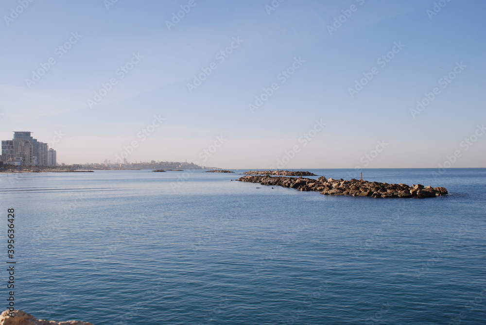Breakwater to the sea and the city in the distance.
Mediterranean Sea, sunny day. A stone breakwater in calm water, against the backdrop of a cityscape. Bright sky.