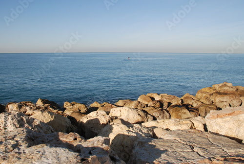 Blue sea stones and clear sky. Mediterranean Sea, sunny day. In the foreground are textured stones illuminated by the sun. Calm sea with blue water. Surfer at sea. Bright clear sky.