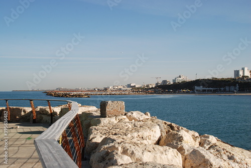 Rectangular stone on a stone pier. Stone pier fenced off with stones and railings. Mediterranean sea, sunny day. The sea is almost calm. In the distance, the silhouette of the city. © Andrew_Swarga