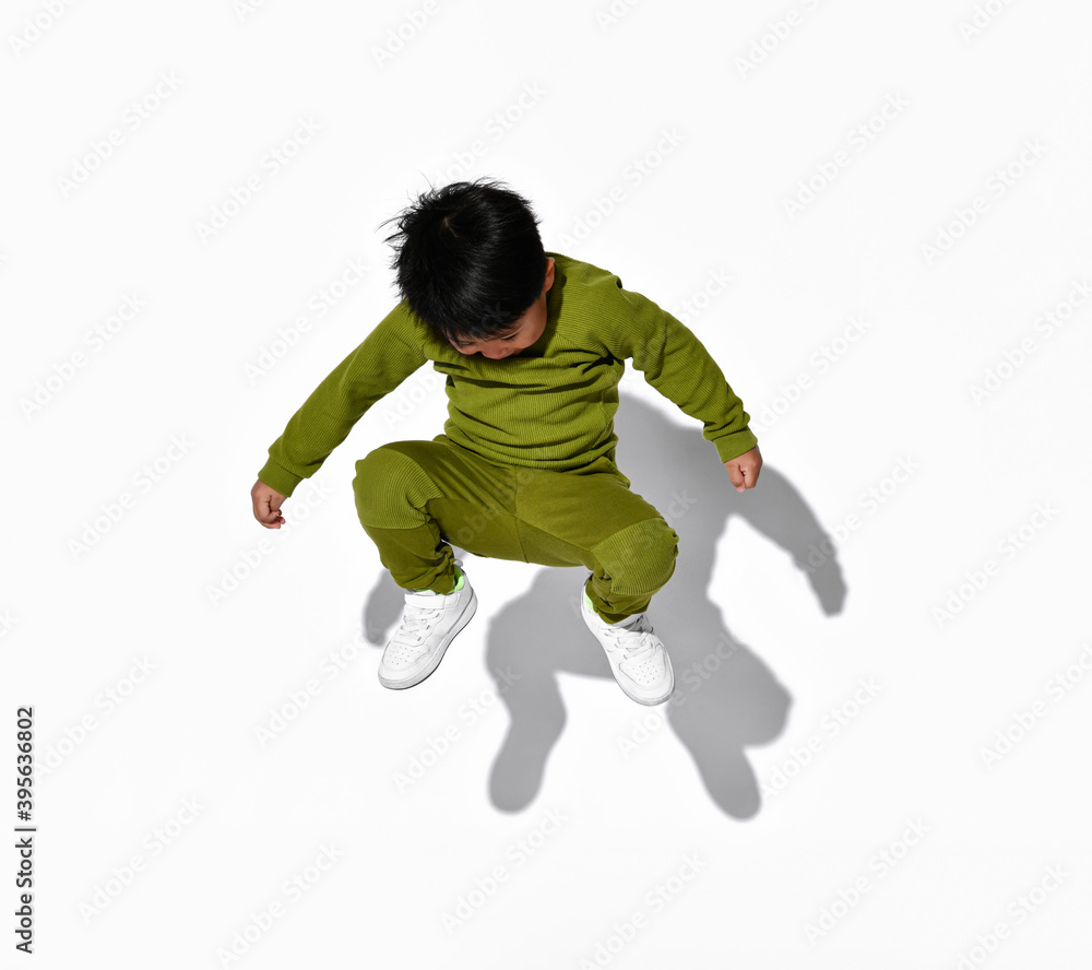 Faceless dark-haired little boy jumps on the background of a white wall in the studio. Child is dressed in a stylish green sports suit and white sneakers. Faceless dark-haired little boy jumps on the background of a white wall in the studio. Child is dressed in a stylish green sports suit and white sneakers.
