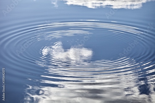 Beautiful glowing rings on blue water surface close-up. Shiny circles pattern with highlights and abstract ripples on calm water surface. White clouds and blue sky reflections on the clear water. 