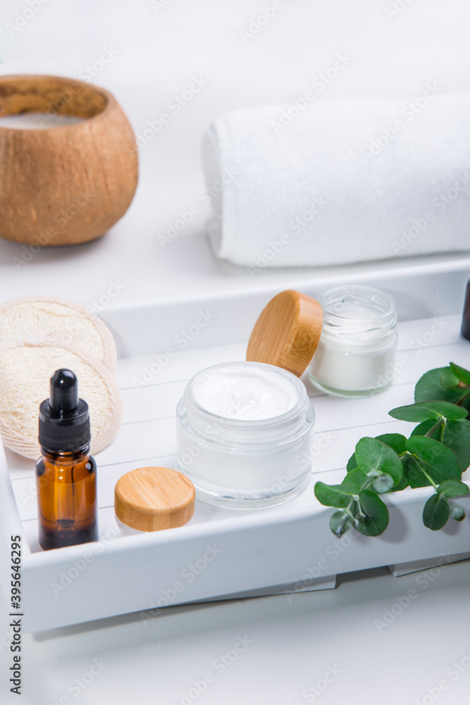 Herbal cosmetic creams in opened containers, fresh eucalyptus, loofah pads and cosmetic natural oil serum on a white wooden tray. Set for face self-care and DIY home eco-friendly hydration. Vertical. Herbal cosmetic creams in opened containers, fresh eucalyptus, loofah pads and cosmetic natural oil serum on a white wooden tray. Set for face self-care and DIY home eco-friendly hydration. Vertical.