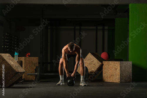 Full-length photo of a handsome man with a naked torso exercising with a kettlebell on a dark background