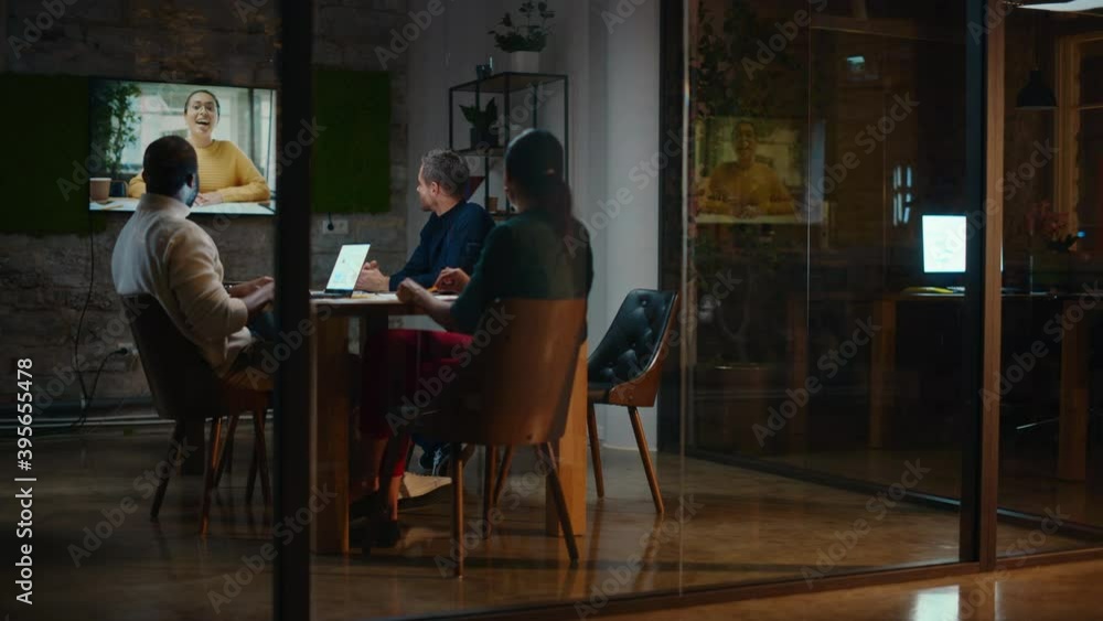 Creative Team is Having a Conference Video Call in Meeting Room Behind Glass Walls in an Office. Colleagues Sit Behind Table and Discuss Business Opportunities with Colleague on Big TV Screen.