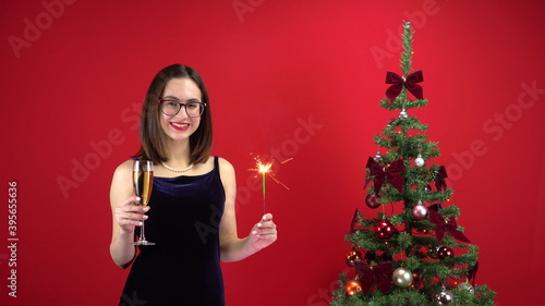 A young woman stands with a glass of champagne and a sparkler near the Christmas tree on a red background. A girl with glasses and an evening dress.