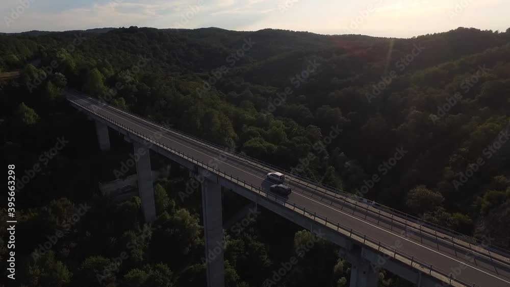 Aerial view of a long and high bridge. Two cars are moving at a moderate speed in an area with many trees and a small river. Aerial view of a long and high bridge. Two cars are moving at a moderate speed in an area with many trees and a small river.