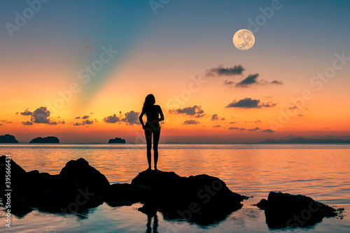 Silhouette of happy young woman standing by the sea on the rocks and looking at the colorful sunset and the full moon 