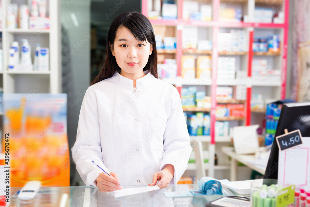 Diligent friendly smiling pleasant chinese woman pharmacist keeps track of drugs in interior of pharmacy.