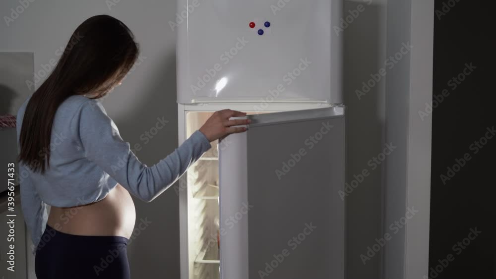 view from the back, a pregnant woman comes to the refrigerator at night and opens the door. Copy space view from the back, a pregnant woman comes to the refrigerator at night and opens the door. Copy space