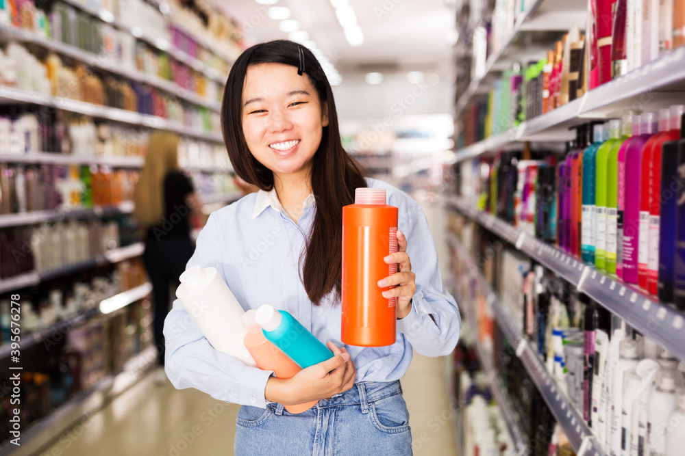 Positive Asian girl holding plastic bottles of shampoo and hair balms in cosmetics store