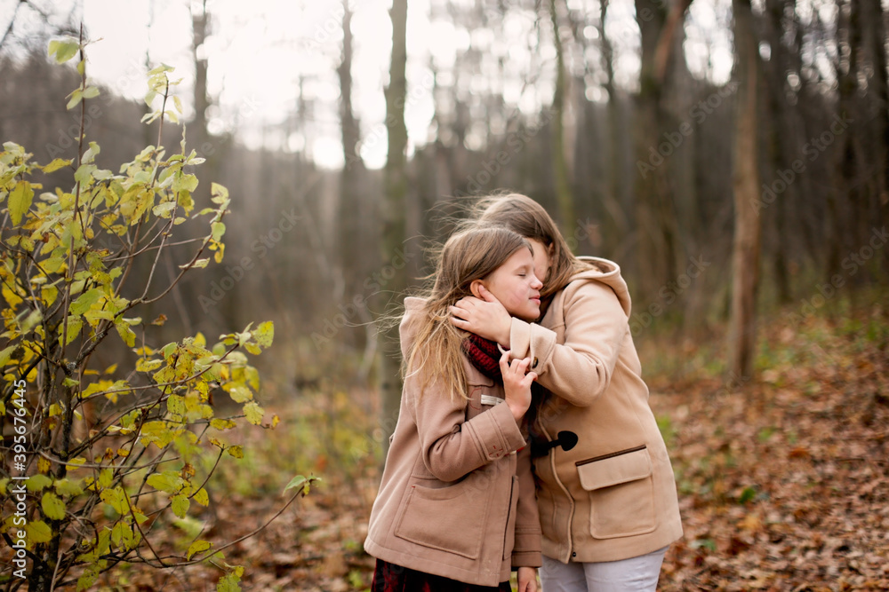 Caucasian children sisters walk together in the park in the forest, girlfriends children hold hands and hug for a walk, girly secrets, autumn walks in warm coats Caucasian children sisters walk together in the park in the forest, girlfriends children hold hands and hug for a walk, girly secrets, autumn walks in warm coats