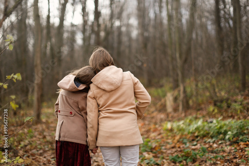 Caucasian children sisters walk together in the park in the forest along the path, girlfriends children hold hands and hug on a walk, view from the back, girly secrets, autumn walks in warm coats