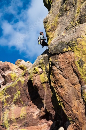 Climber repelling down from a tall multicolored rock face in Boulder Colorado. The climber is using climbing equipment to descend from one of the Flatirons
