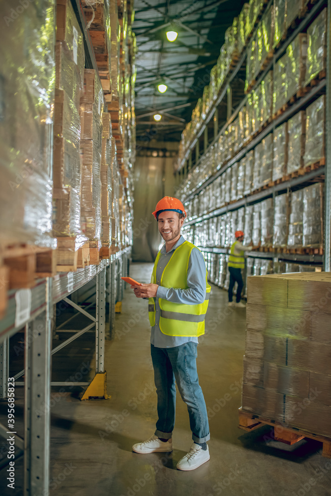 Young warehouse worker in helmet standing near containers and holding a tablet Young warehouse worker in helmet standing near containers and holding a tablet
