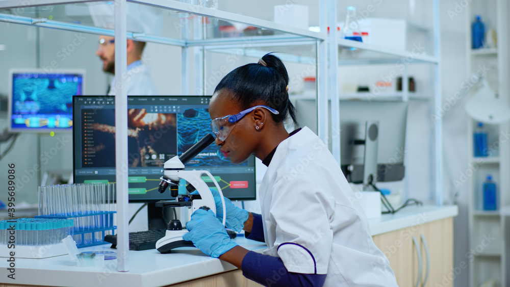 African scientist checking sample of virus using microscope in modern lab. Multiethnic team examining vaccine evolution using high tech for scientific research of treatment development against covid19 - obrazy, fototapety, plakaty 
